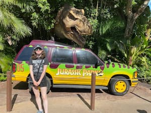 author standing in front of car while T-Rex structure pokes its head out behind them